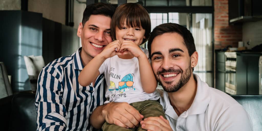 Casal homoafetivo masculino sorridente com seu filho pequeno no colo, representando uma família formada por fertilização in vitro (FIV).