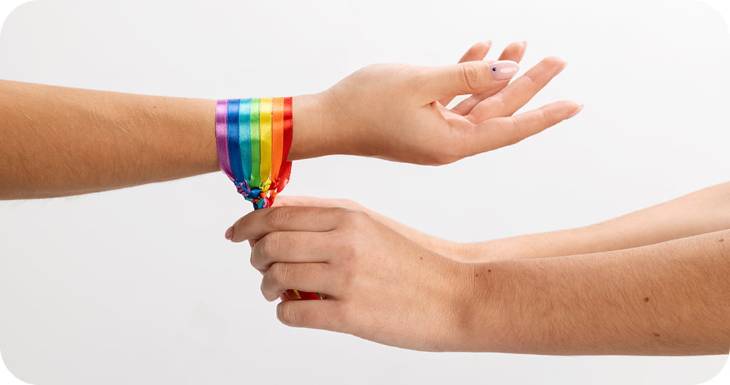 Duas mãos interagindo em um fundo branco. A mão à esquerda está com um laço ou fita nas cores do arco-íris amarrada ao redor do pulso. A mão à direita está segurando ou ajustando essa fita colorida no pulso da outra pessoa. 