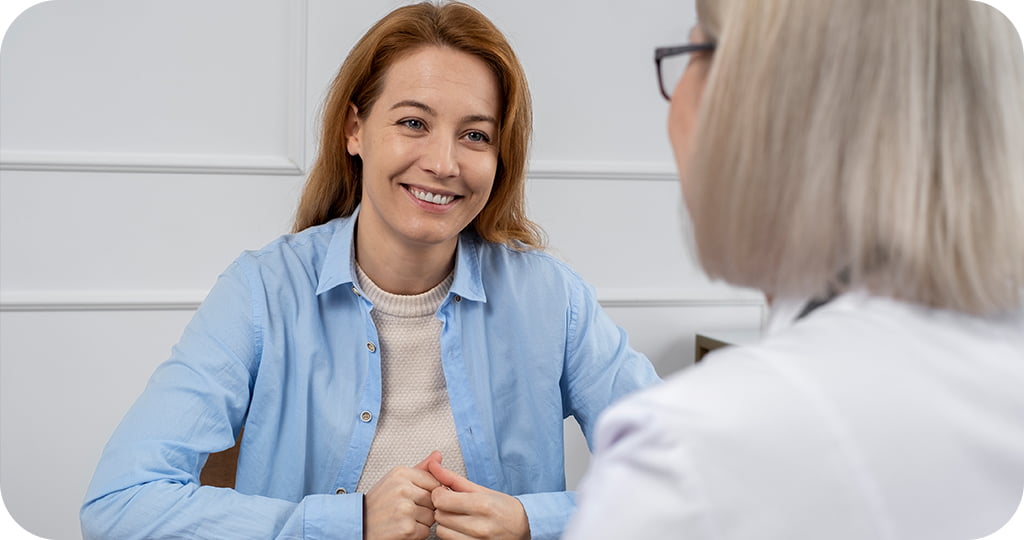 duas pessoas em um ambiente interno, aparentemente em uma consulta médica. A pessoa à esquerda está vestindo uma camisa azul clara sobre uma blusa bege, e parece estar sorrindo enquanto conversa com a outra pessoa.
