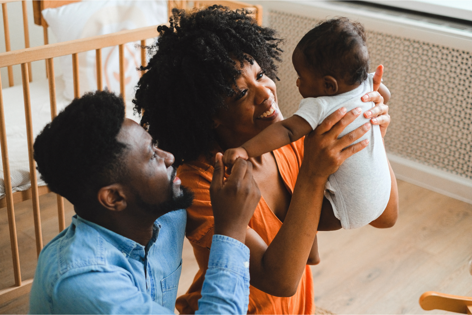 A imagem mostra uma família composta por três pessoas: dois adultos e um bebê. Os adultos parecem ser um homem e uma mulher. O homem, que está ajoelhado ou agachado, veste uma camisa jeans azul e tem barba. A mulher está sentada, usando um vestido laranja, e segura o bebê, que está vestindo um body branco. Ambos os adultos estão sorrindo e olhando para o bebê, demonstrando carinho e felicidade.