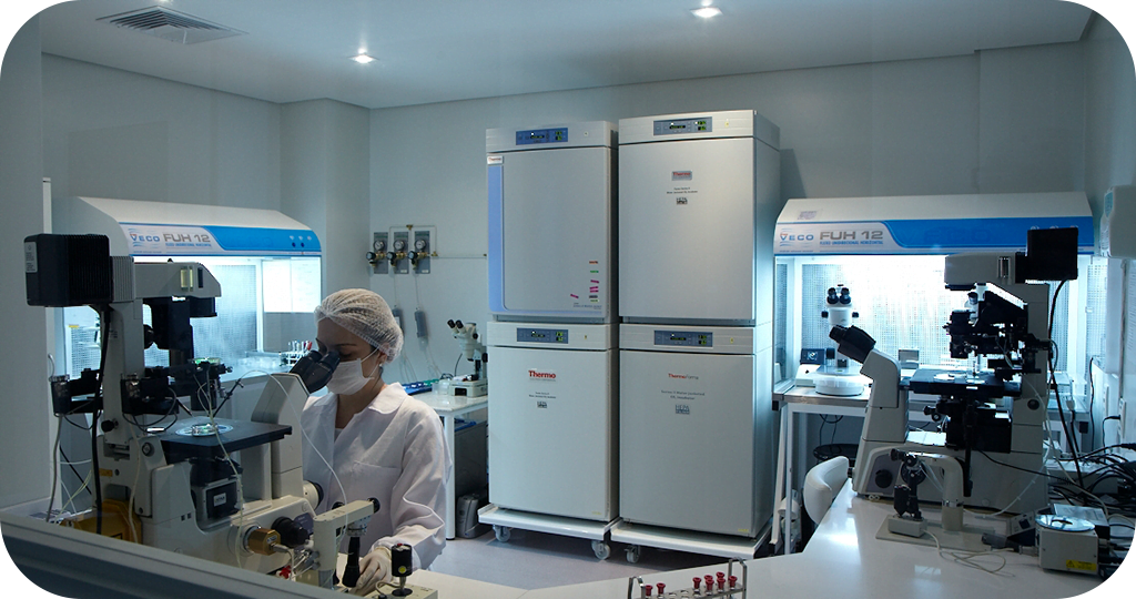 A imagem mostra um laboratório moderno e bem equipado. No centro, há uma pessoa usando um jaleco branco, touca e máscara, trabalhando em um microscópio. O ambiente é limpo e bem iluminado, com vários equipamentos de laboratório visíveis ao fundo, incluindo dois grandes refrigeradores de laboratório da marca Thermo e duas cabines de fluxo laminar da marca Veco. À direita e à esquerda da pessoa, há mais microscópios e outros instrumentos científicos sobre as bancadas. O laboratório parece estar focado em pesquisas biológicas ou médicas, possivelmente em um campo como a medicina reprodutiva ou a biotecnologia.