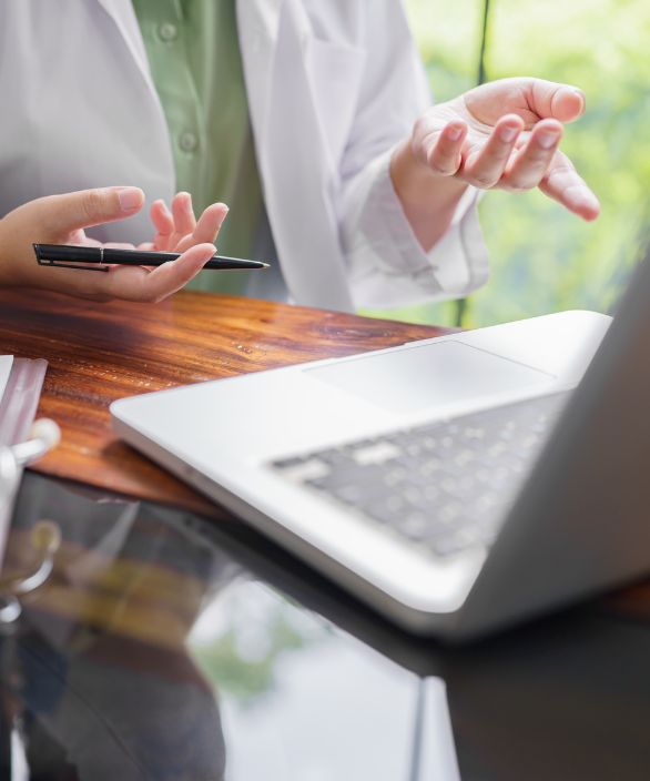 Médico ou médica com jaleco branco gesticulando durante uma videoconferência em frente a um notebook sobre a mesa.