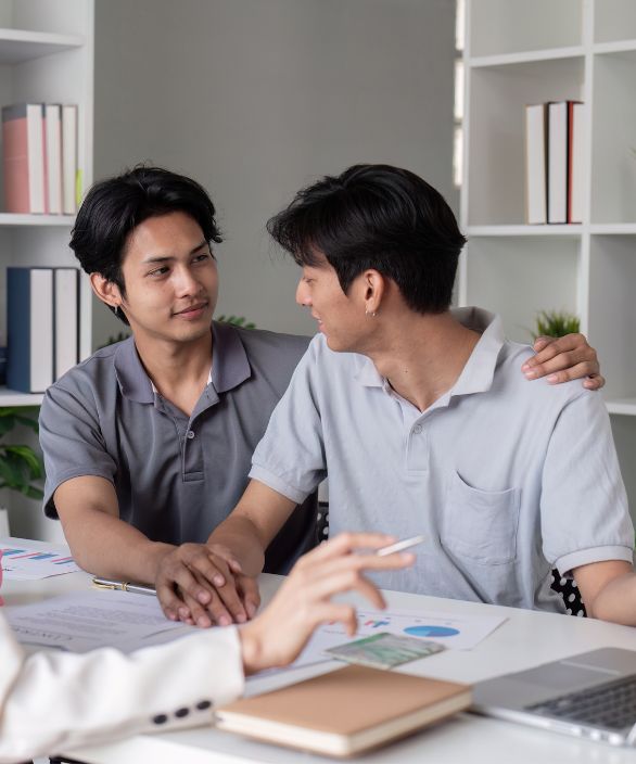 Casal homoafetivo masculino sentado em uma mesa de atendimento, trocando olhares afetuosos enquanto seguram as mãos, durante uma consulta em ambiente profissional. Imagem associada à Nilo Frantz Medicina Reprodutiva.