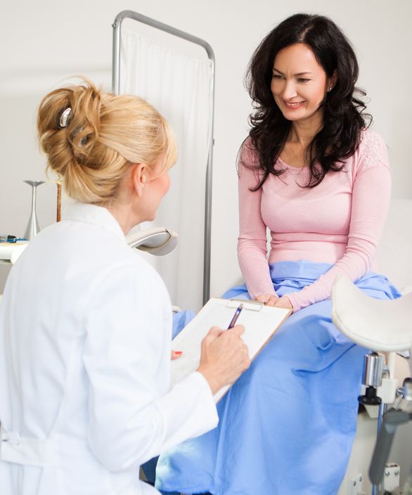 Mulher sorridente sentada em uma cadeira ginecológica durante consulta médica, conversando com uma profissional de saúde que segura uma prancheta. Imagem relacionada à Nilo Frantz Medicina Reprodutiva.