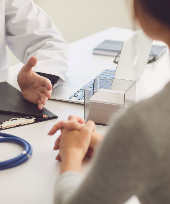 Pessoa em consulta médica, sentada à frente de um profissional da saúde, com as mãos entrelaçadas sobre a mesa. Estetoscópio, laptop e materiais de trabalho também estão visíveis, sugerindo uma conversa atenciosa e profissional.