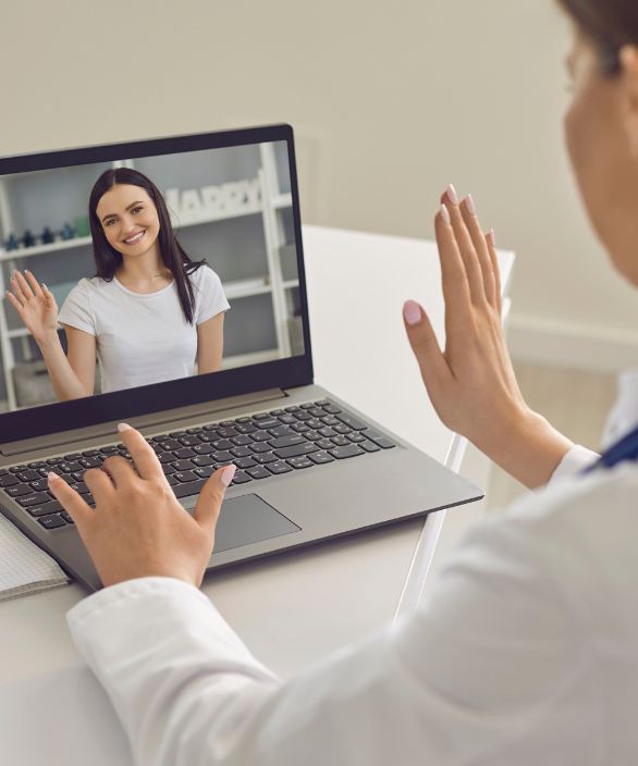 Profissional de saúde realiza videochamada com paciente, ambas sorrindo e acenando uma para a outra por meio da tela do notebook.