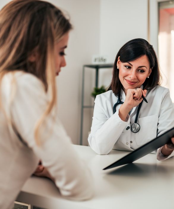 Médica sorridente em consulta com paciente, segurando um prontuário e explicando informações de forma acolhedora. Imagem associada à Nilo Frantz Medicina Reprodutiva.