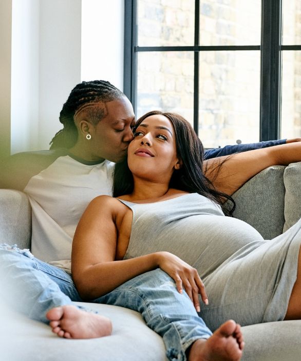 Casal homoafetivo feminino sentado no sofá, em um momento íntimo e afetuoso. Uma das mulheres está grávida, sendo acarinhada com um beijo na testa pela parceira. Imagem associada à Nilo Frantz Medicina Reprodutiva.