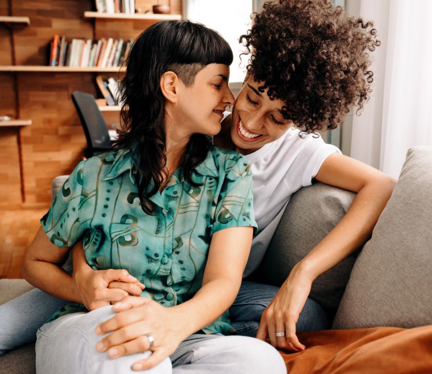 Casal sorridente e carinhoso sentado em um sofá, representando afeto e união, com foco na diversidade e no amor. Ambas usam anéis nas mãos esquerdas, sugerindo compromisso. Imagem relacionada à Nilo Frantz Medicina Reprodutiva.