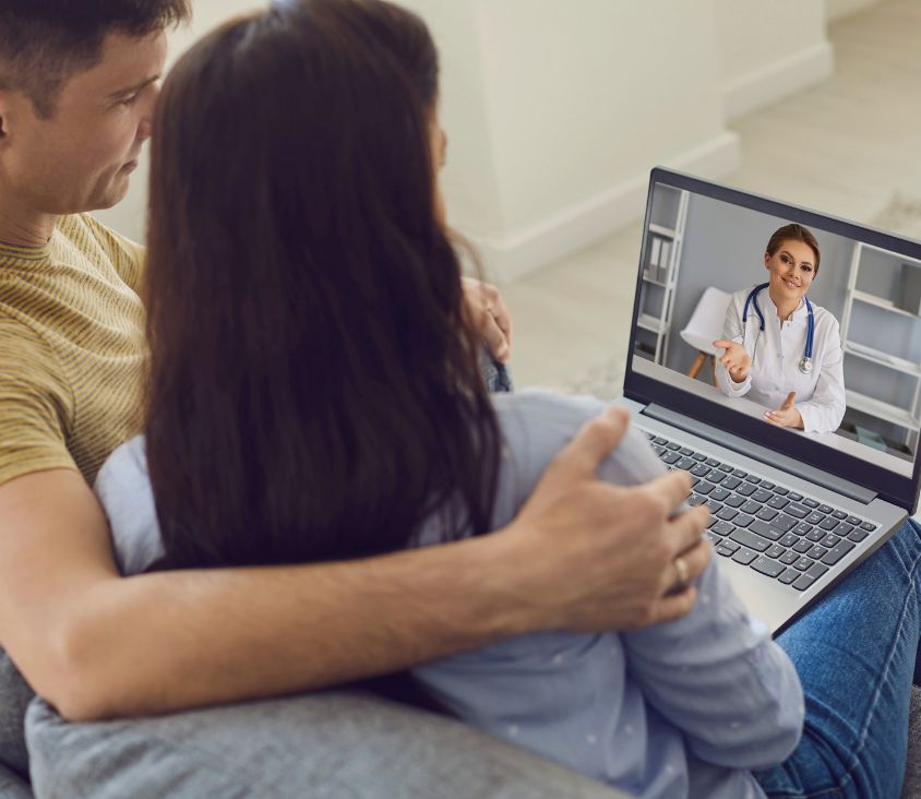 Casal sentado em casa participando de uma consulta médica online com uma médica em vídeo chamada, visível na tela do notebook.