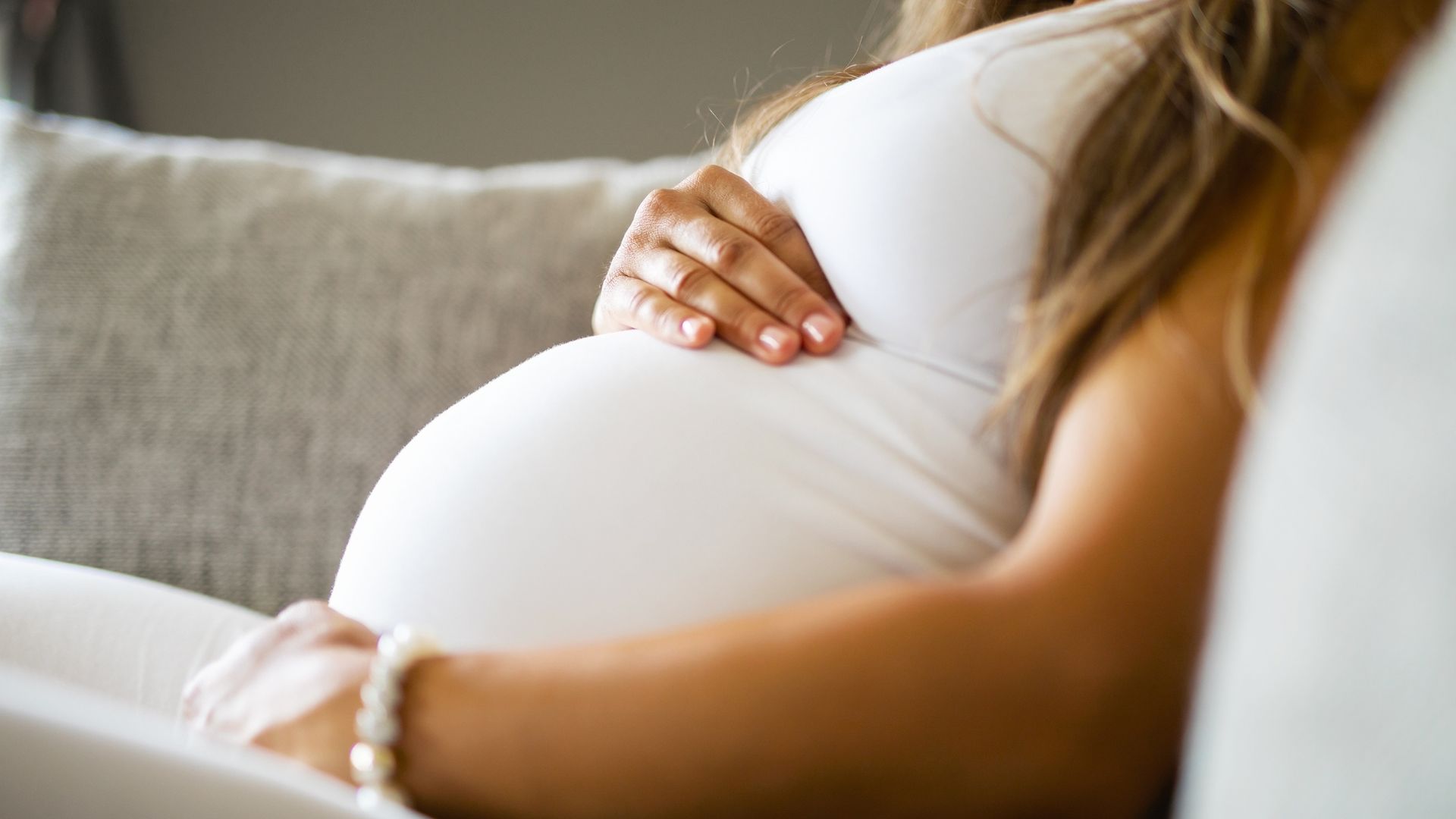 Mulher grávida descansando no sofá, com as mãos apoiadas sobre a barriga, representando acolhimento, maternidade e cuidado durante a gestação.